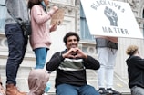 People participate in a demonstration with signs supporting the Black Lives Matter movement. A young man sits in the foreground making a heart gesture with his hands, surrounded by others standing and holding signs.