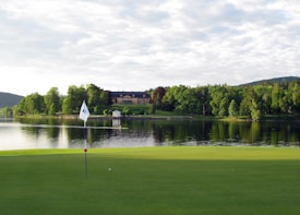 A serene golf course with a green foreground featuring a flagstick and cup. In the background, there's a calm lake reflecting the surrounding trees and a large building with a pitched roof. The scene is lush with greenery and appears peaceful under a partly cloudy sky.