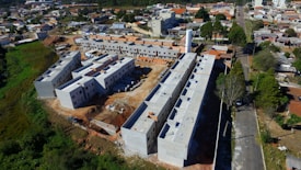 Aerial view of a construction site featuring multiple partially constructed buildings with a combination of flat and sloped roofs. The site is surrounded by a mix of residential houses and green areas. There are construction materials scattered around, and the roads nearby are bordered by trees and small residential properties.