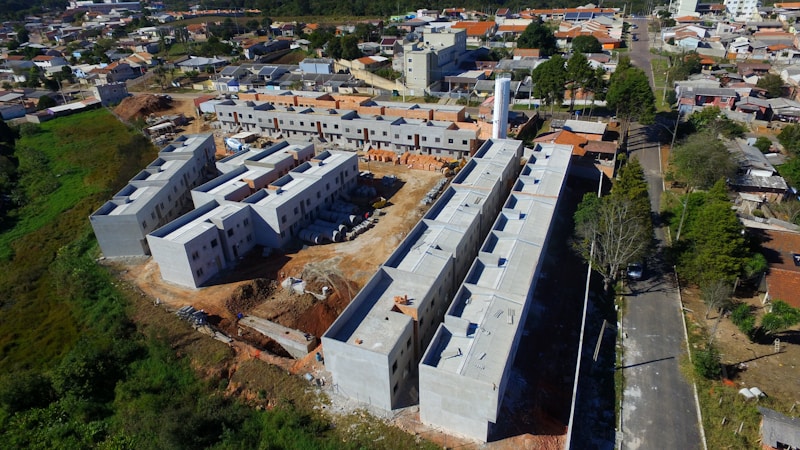 Aerial view of a construction site featuring multiple partially constructed buildings with a combination of flat and sloped roofs. The site is surrounded by a mix of residential houses and green areas. There are construction materials scattered around, and the roads nearby are bordered by trees and small residential properties.
