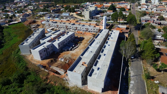 Aerial view of a construction site featuring multiple partially constructed buildings with a combination of flat and sloped roofs. The site is surrounded by a mix of residential houses and green areas. There are construction materials scattered around, and the roads nearby are bordered by trees and small residential properties.