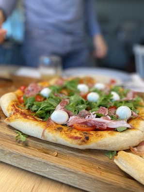 A freshly baked pizza topped with arugula, cherry tomatoes, mozzarella balls, and slices of prosciutto on a wooden board. A person wearing a blue long-sleeve shirt is blurred in the background, suggesting activity in a kitchen or dining area.