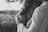 A black and white photograph featuring a close embrace between two people. One person is holding the other gently, and there is an intimate, tender interaction. The focus is on their hands and the soft expression of the person being held. The background is blurred, suggesting an outdoor setting.