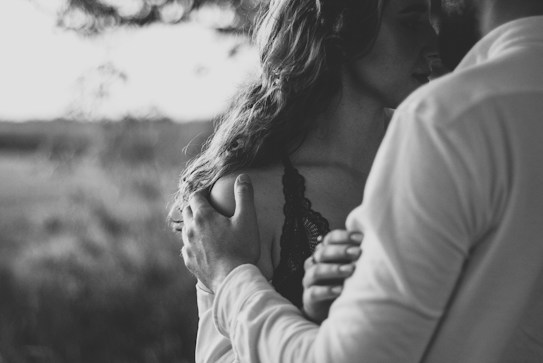 A black and white photograph featuring a close embrace between two people. One person is holding the other gently, and there is an intimate, tender interaction. The focus is on their hands and the soft expression of the person being held. The background is blurred, suggesting an outdoor setting.