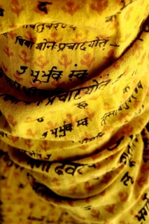 A close-up view of yellow fabric with black script text printed on it. The fabric appears to be folded or layered, creating shadows and depth. There are subtle reddish floral patterns interspersed with the text.