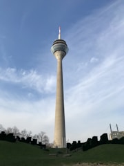 A tall telecommunications tower rises against a clear blue sky, surrounded by a grassy area with scattered trees and foliage. A bridge is visible in the background on the right side.