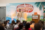 A man is standing on a stage in front of a backdrop with the text 'Social Media Marketing World'. He is wearing a patterned shirt and white pants, gesturing as he speaks. There is a podium with a microphone and two books on it. An audience is seated and listening attentively.