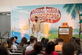 A man is standing on a stage in front of a backdrop with the text 'Social Media Marketing World'. He is wearing a patterned shirt and white pants, gesturing as he speaks. There is a podium with a microphone and two books on it. An audience is seated and listening attentively.