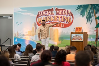 A man is standing on a stage in front of a backdrop with the text 'Social Media Marketing World'. He is wearing a patterned shirt and white pants, gesturing as he speaks. There is a podium with a microphone and two books on it. An audience is seated and listening attentively.
