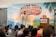 A man is standing on a stage in front of a backdrop with the text 'Social Media Marketing World'. He is wearing a patterned shirt and white pants, gesturing as he speaks. There is a podium with a microphone and two books on it. An audience is seated and listening attentively.
