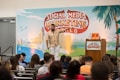 A man is standing on a stage in front of a backdrop with the text 'Social Media Marketing World'. He is wearing a patterned shirt and white pants, gesturing as he speaks. There is a podium with a microphone and two books on it. An audience is seated and listening attentively.