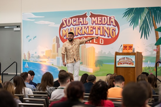 A man is standing on a stage in front of a backdrop with the text 'Social Media Marketing World'. He is wearing a patterned shirt and white pants, gesturing as he speaks. There is a podium with a microphone and two books on it. An audience is seated and listening attentively.