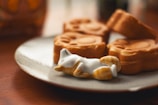 A ceramic figurine of a reclining animal, possibly a cat, is positioned on a plate next to three brown pastries shaped like animal faces. The scene is set on a wooden table, and the lighting creates a warm and cozy atmosphere.