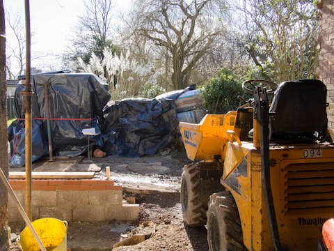 A construction site with a small brick wall in progress. A yellow dumper truck is parked nearby, and there are large bags covered with blue tarpaulin. The area is surrounded by trees and bushes, and there is a bright sunny environment.