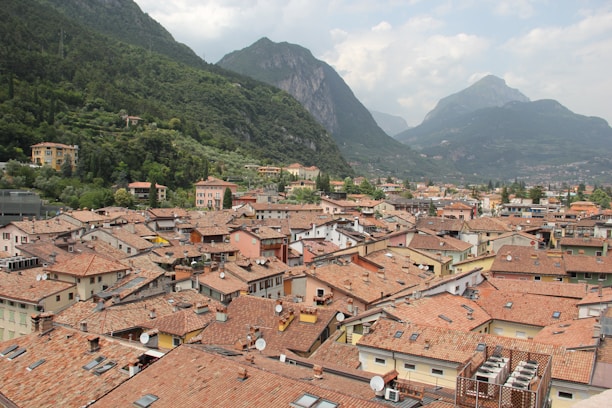 A picturesque view of a European town with terracotta roofed houses. The town is nestled in a valley surrounded by lush green mountains. The sky is partly cloudy, creating a serene atmosphere. Various buildings show distinct architectural styles typical of a traditional European setting.