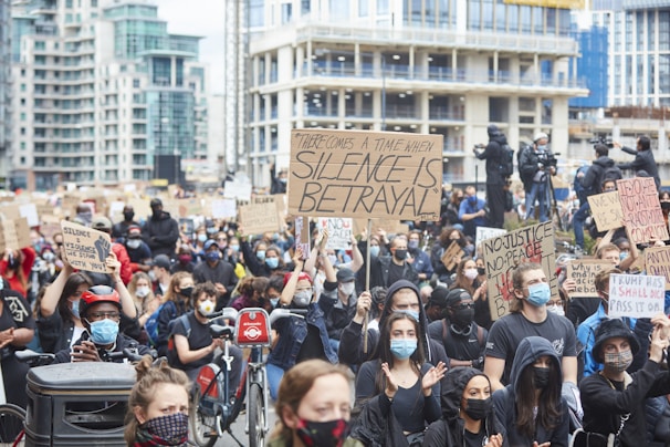 A large crowd of people participates in a protest in an urban setting, holding various signs with messages advocating for justice and change. Most individuals are wearing face masks, and some are sitting on bikes. The background features tall buildings and additional people capturing the event with cameras.