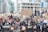 A large crowd of people participates in a protest in an urban setting, holding various signs with messages advocating for justice and change. Most individuals are wearing face masks, and some are sitting on bikes. The background features tall buildings and additional people capturing the event with cameras.