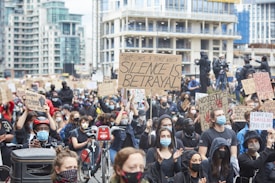 A large crowd of people participates in a protest in an urban setting, holding various signs with messages advocating for justice and change. Most individuals are wearing face masks, and some are sitting on bikes. The background features tall buildings and additional people capturing the event with cameras.