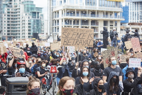 A large crowd of people participates in a protest in an urban setting, holding various signs with messages advocating for justice and change. Most individuals are wearing face masks, and some are sitting on bikes. The background features tall buildings and additional people capturing the event with cameras.
