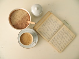 A flat lay image features a cup filled with frothy coffee, placed on a saucer, a bowl containing a mixture of light and dark brown ingredients with a wooden spoon, a white ceramic sugar jar with a lid, and an open notebook filled with handwritten entries, all set against a light background.