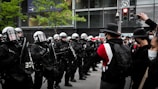 A group of police officers in riot gear stand in formation facing a crowd of protesters. The officers are wearing helmets and holding shields, while the protesters are wearing masks and some are carrying signs. There is a traffic light in the background, along with some green trees and a modern building facade.