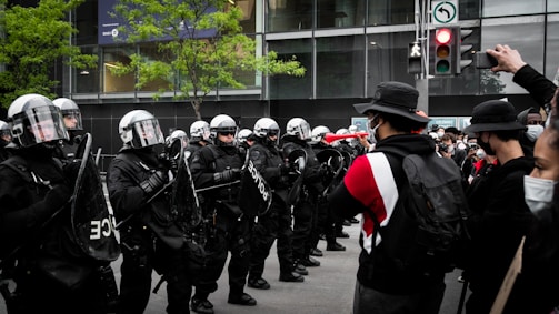 A group of police officers in riot gear stand in formation facing a crowd of protesters. The officers are wearing helmets and holding shields, while the protesters are wearing masks and some are carrying signs. There is a traffic light in the background, along with some green trees and a modern building facade.