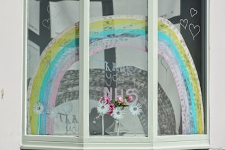 A large rainbow graphic is drawn on a window, featuring vibrant colors in stripes. Below the rainbow, there is a vase with pink and white flowers, and several white daisy-like drawings. The words 'Thank You NHS' are written in the middle of the window. Several heart shapes are doodled around the rainbow, creating a decorative and uplifting display.