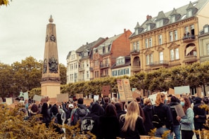 A crowd of people is gathered in front of a historic, European-style building, holding up signs and banners in a protest or demonstration. The scene includes a tall, ornate monument, resembling an obelisk, situated among trees.