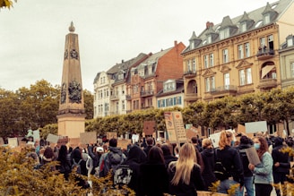 A crowd of people is gathered in front of a historic, European-style building, holding up signs and banners in a protest or demonstration. The scene includes a tall, ornate monument, resembling an obelisk, situated among trees.