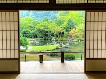 A traditional Japanese-style room opens up to a serene garden with lush green trees, colorful foliage, a reflective pond, and strategically placed rocks. The paper sliding doors frame a view that blends nature with architecture.