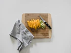 Hand holding a bamboo cutting board next to a set of sharp knives and fresh vegetables on a clean countertop.