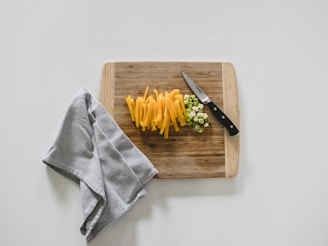 A wooden cutting board is placed on a light surface with sliced yellow bell peppers and chopped green onions neatly organized. A knife with a black handle lies next to the vegetables. Beside the cutting board, a grey textured cloth is casually draped.