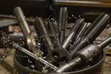 A collection of various metal tools, including taps and drill bits, are placed upright in a container. The tools have a metallic sheen and are surrounded by curly metal shavings, suggesting a workshop setting.