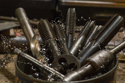 A collection of various metal tools, including taps and drill bits, are placed upright in a container. The tools have a metallic sheen and are surrounded by curly metal shavings, suggesting a workshop setting.