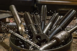 A collection of various metal tools, including taps and drill bits, are placed upright in a container. The tools have a metallic sheen and are surrounded by curly metal shavings, suggesting a workshop setting.