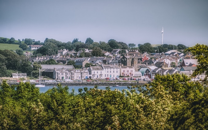 A quaint coastal town with a collection of houses and buildings nestled among lush green trees. A wind turbine is visible in the distance, indicating an interest in renewable energy. The town is bordered by a body of water in the foreground and rolling hills in the background.