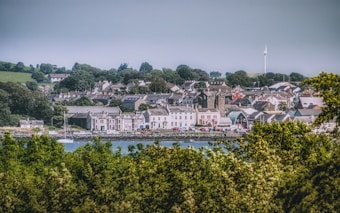 A quaint coastal town with a collection of houses and buildings nestled among lush green trees. A wind turbine is visible in the distance, indicating an interest in renewable energy. The town is bordered by a body of water in the foreground and rolling hills in the background.