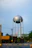 A tall metal water tower stands prominently in the foreground, featuring a spherical tank on stilts. Below the tower, there is a red-brick building surrounded by trees and power lines. A faint rainbow is visible in the background, contrasting with the overcast sky.