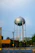 A tall metal water tower stands prominently in the foreground, featuring a spherical tank on stilts. Below the tower, there is a red-brick building surrounded by trees and power lines. A faint rainbow is visible in the background, contrasting with the overcast sky.