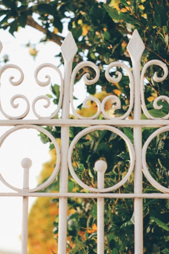 A decorative white wrought iron fence with intricate spirals and pointed tips. Behind the fence, lush green foliage is visible, adding a natural backdrop.