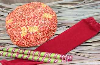 A red and orange patterned turban cap is placed on a background of dried leaves alongside a pair of red socks and multiple green and pink decorated sticks.