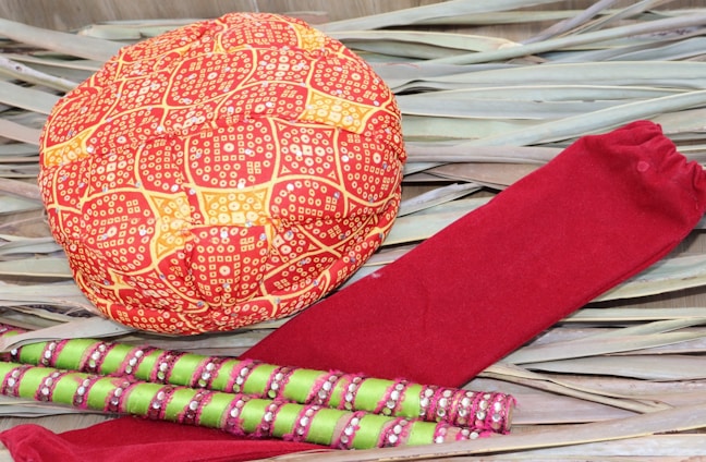 A red and orange patterned turban cap is placed on a background of dried leaves alongside a pair of red socks and multiple green and pink decorated sticks.