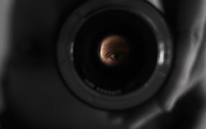 An eye is visible through the lens of a camera, with the surrounding area in shadow, suggesting a theme of observation or photography.