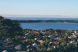 A scenic view overlooks a residential area with various houses spread out among trees. In the background, a large body of water stretches into the horizon under a clear blue sky. The foreground features lush greenery, and a tree is visible on the left side.