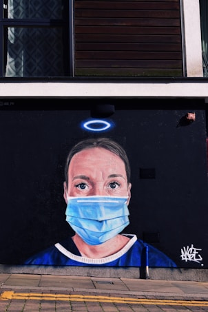 A mural painted on the side of a building depicts a person wearing a blue medical mask and blue scrubs. Above their head is a halo. The background is dark, highlighting the portrait. The mural is located on a street with a window above and a sidewalk in front.
