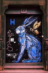 A mural painted on a shutter door features a stylized blue hare accompanied by a large yellow insect, possibly a bee. The artwork includes elaborate line work and graffiti-style elements, set against a black background. Above, a sign reads '22 Lever St' indicating the building's address.