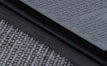 An e-reader device displaying a page of text is partially visible, resting on a textured fabric surface. The focus is on the edge of the device, highlighting its black frame and digital screen with gray text.