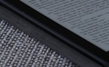 An e-reader device displaying a page of text is partially visible, resting on a textured fabric surface. The focus is on the edge of the device, highlighting its black frame and digital screen with gray text.