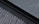 An e-reader device displaying a page of text is partially visible, resting on a textured fabric surface. The focus is on the edge of the device, highlighting its black frame and digital screen with gray text.