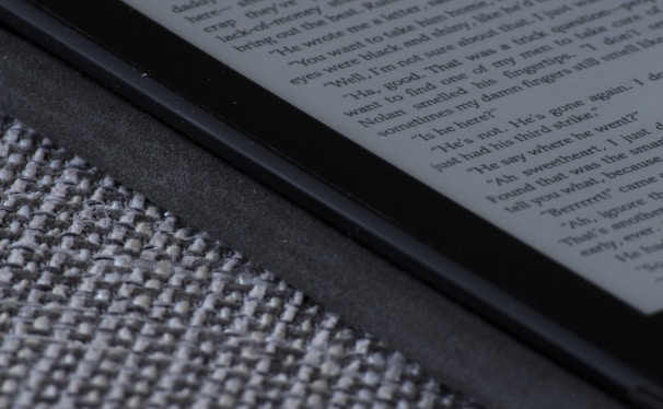 An e-reader device displaying a page of text is partially visible, resting on a textured fabric surface. The focus is on the edge of the device, highlighting its black frame and digital screen with gray text.